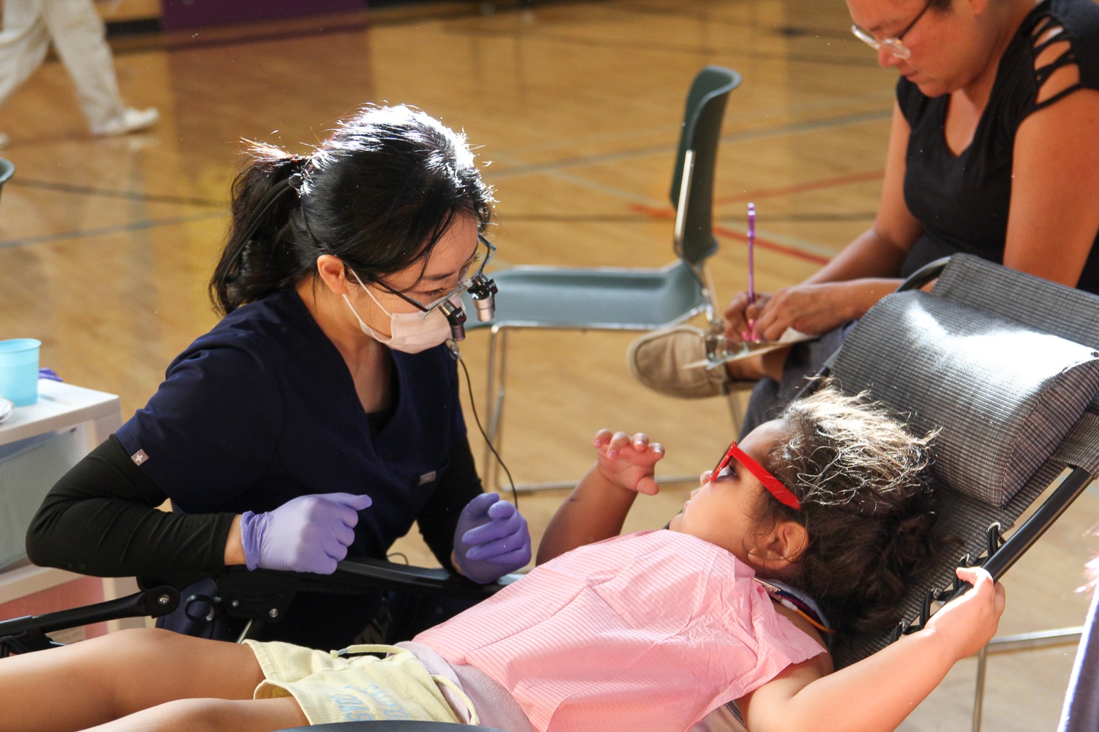 A Wings for Smiles hygienist gently treats a young girl wearing red safety glasses in the Chase community gym