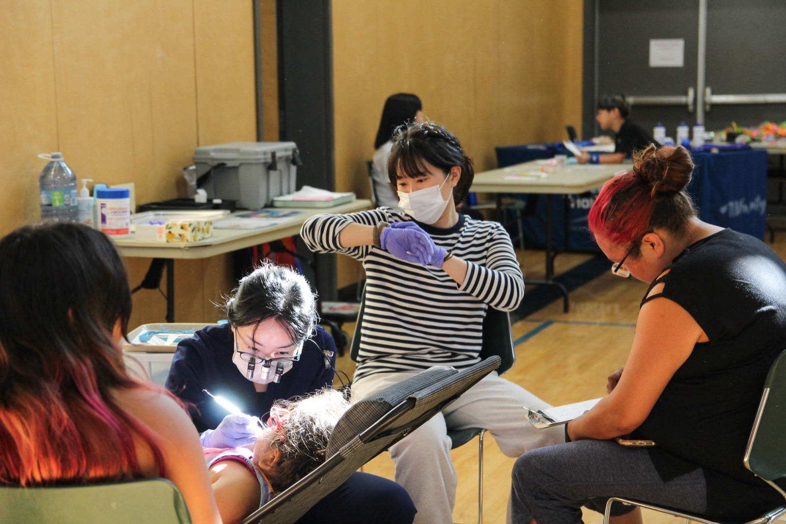 A hygienist with loupes treats a child while volunteers and family look on in the gym