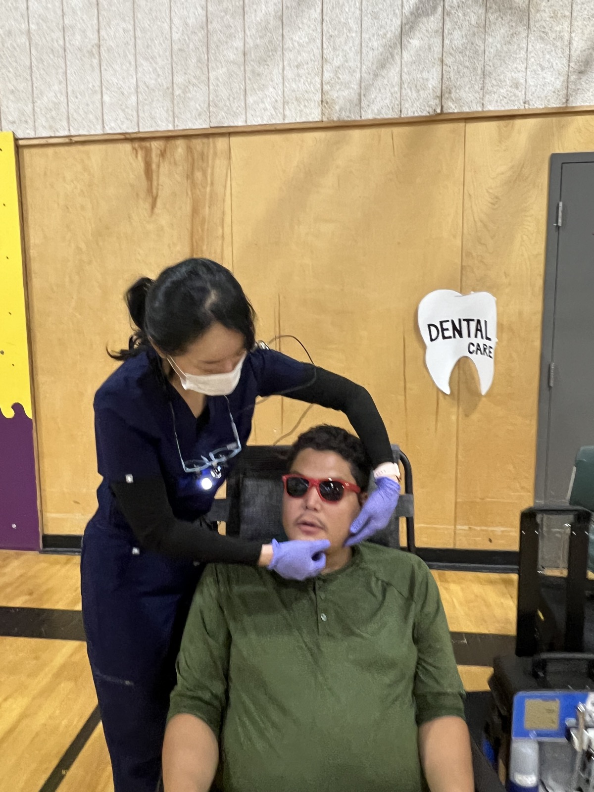 A hygienist examines a young man beneath a hand-made Dental Care tooth sign in the community gym
